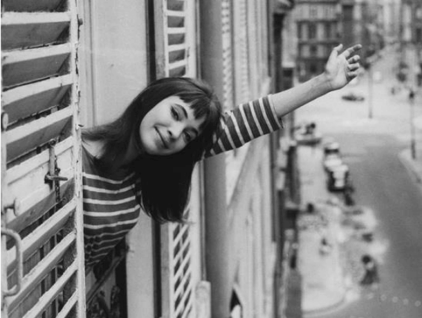 Anna KArina, una de las actrices de la nouvelle vague, asomada a una ventana sonriendo a cámara y saludando con la mano