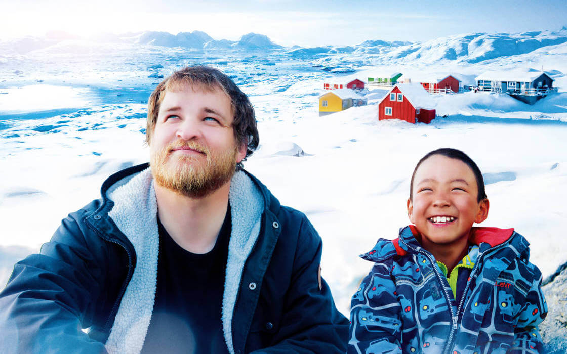 Los principales protagonistas de Un profesor en Groenlandia sonriendo y con el paisaje nevado de la aldea inuit en la que se desarrolla la película