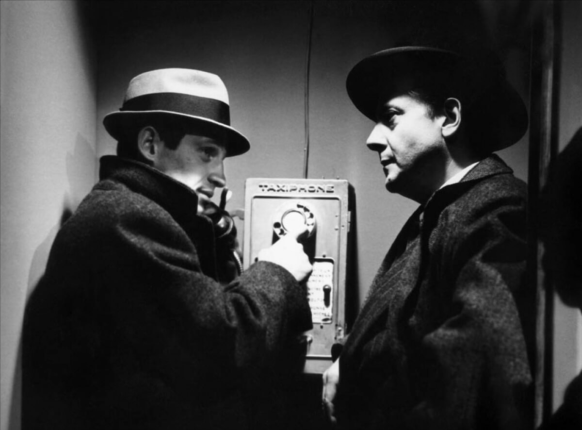 Dos gánsters con la típica imagen del cine polar: sombrero y gabardina, en una cabina de teléfono pública.
